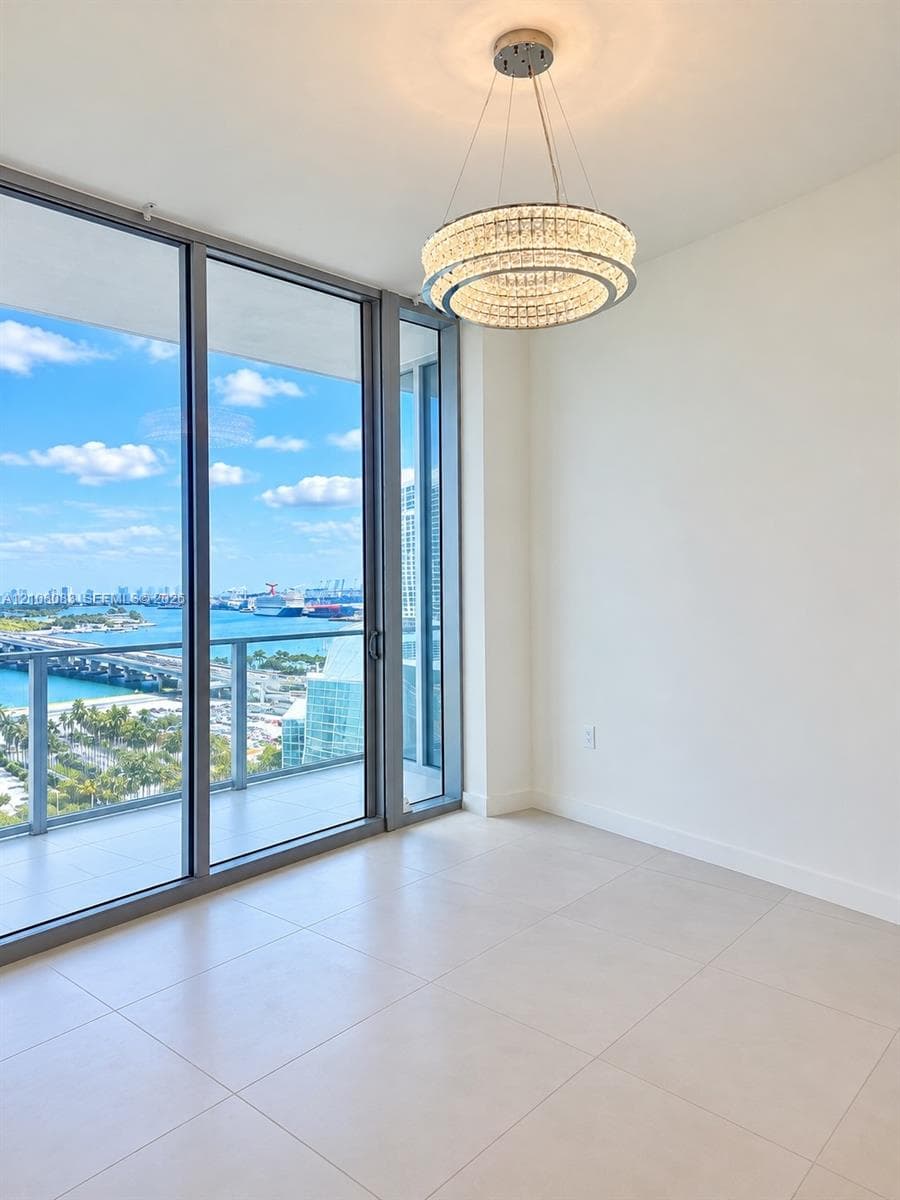 Floor-to-ceiling glass panel windows, wide-format tile flooring, multi-tiered crystal chandelier, and integrated glass balcony railing with water and cityscape backdrop