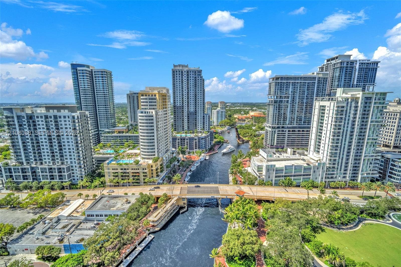 Aerial perspective of a high-density urban corridor featuring a central waterway, bridge infrastructure, mature landscaping, and numerous mid-rise and high-rise structures
