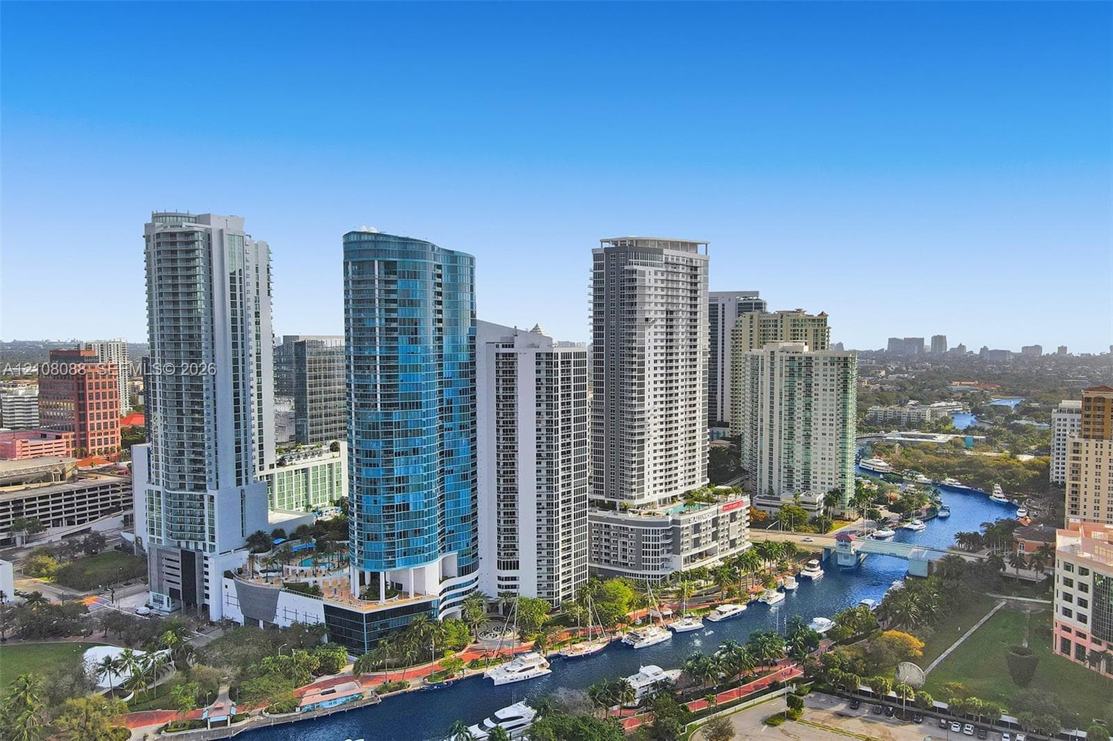 Aerial perspective of a high-rise urban landscape featuring reflective glass facades, multiple waterfront buildings, a navigable waterway, and landscaped green space