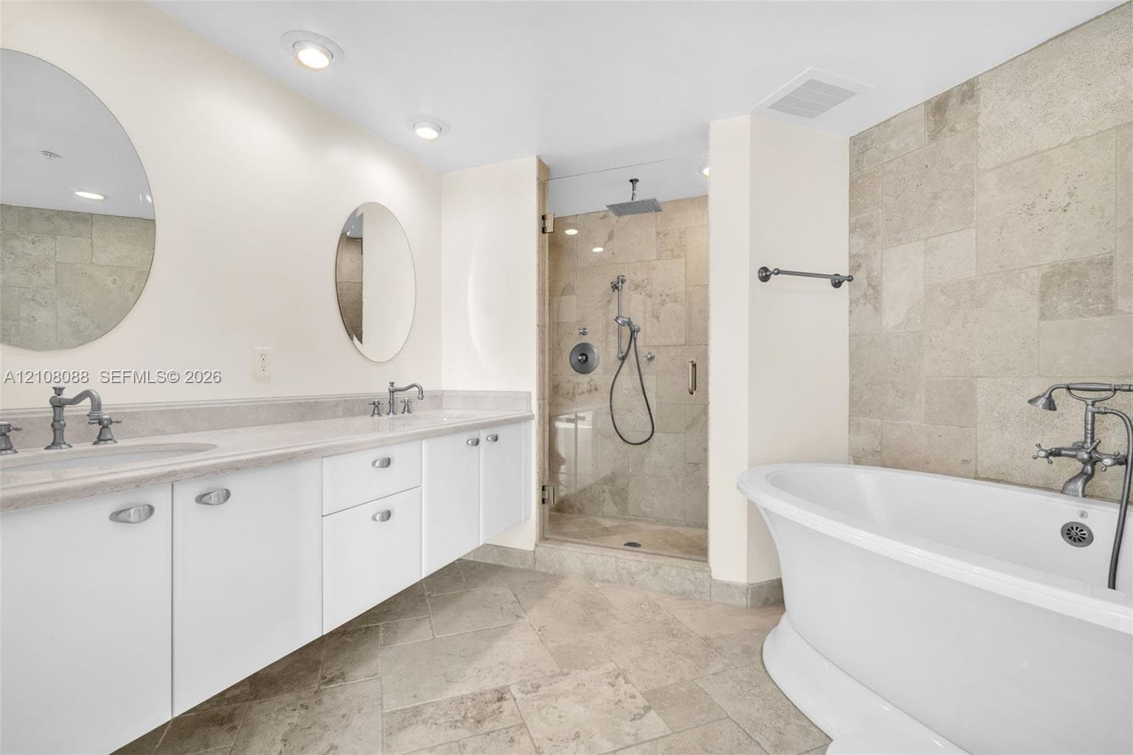 Bathroom featuring a double vanity with stone countertops, a freestanding soaking tub, a glass-enclosed shower with stone tile surround, and recessed lighting