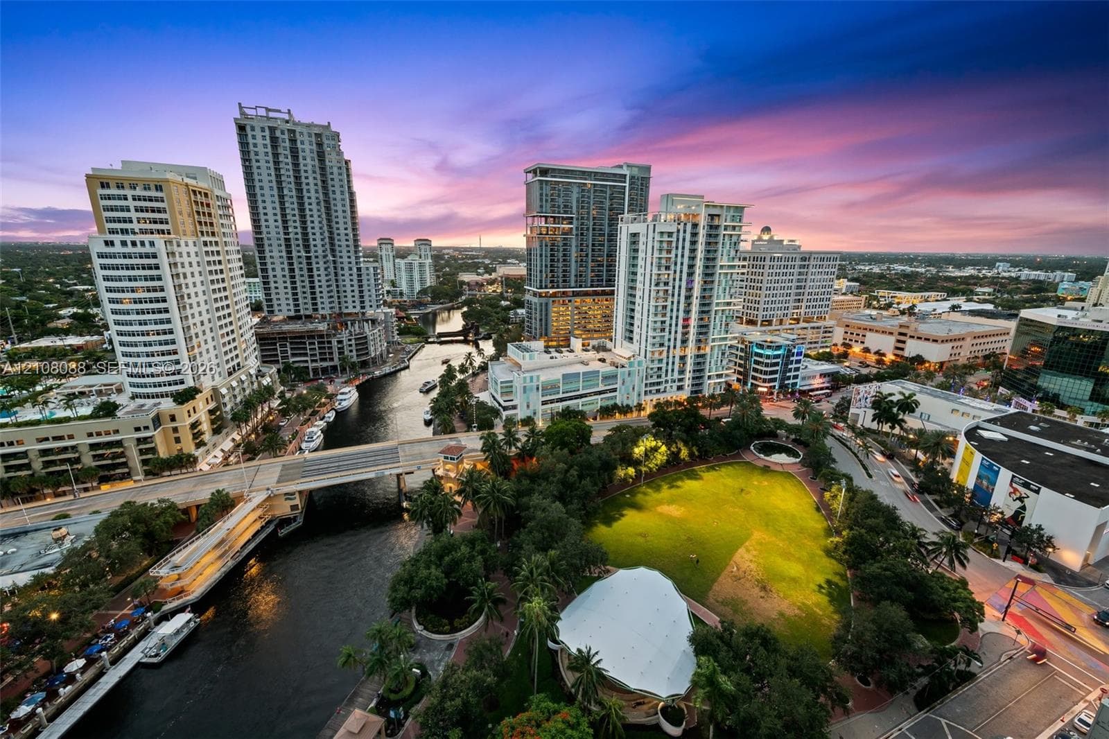 Aerial perspective showcasing a metropolitan skyline, a winding waterway, a central urban park with a structured pavilion, and mature landscaping