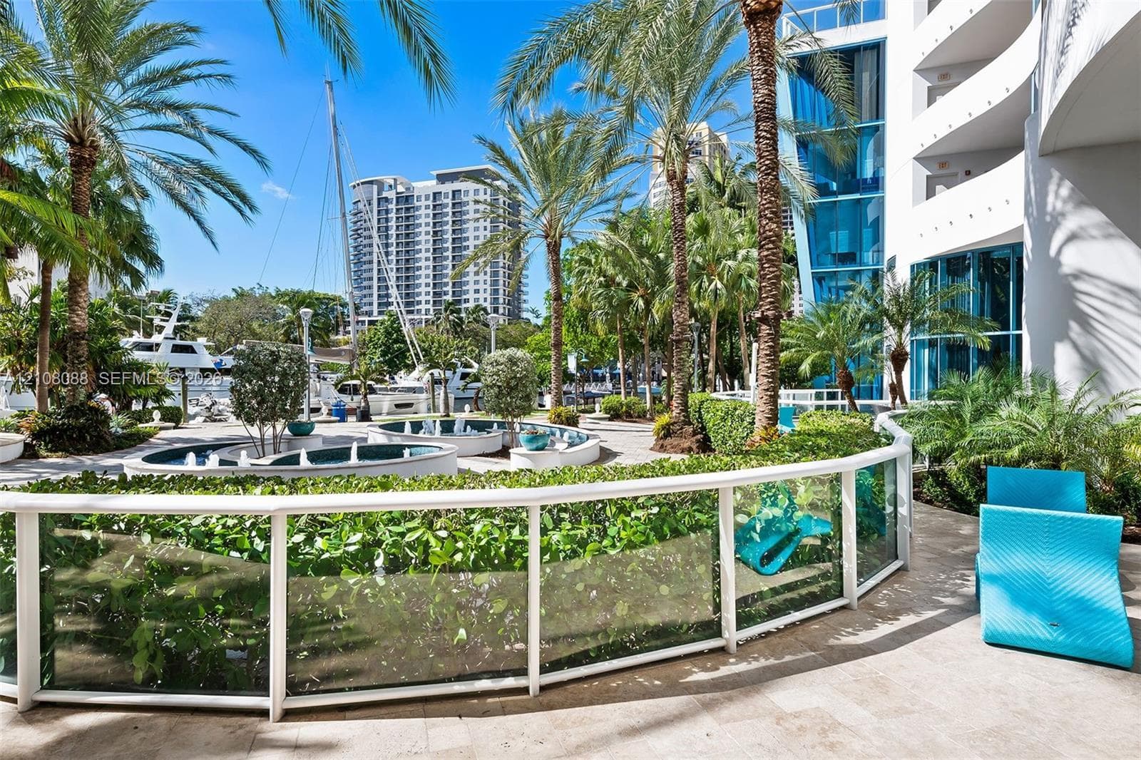 Outdoor terrace featuring stone flooring, glass panel railings, and mature palm trees, complemented by built-in circular fountains and manicured shrubbery