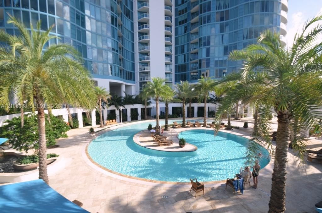 Curved swimming pool with natural stone decking, palm tree landscaping, and a multi-story glass-facade high-rise structure