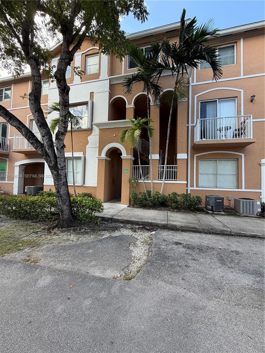 Multi-story exterior featuring stucco siding, arched entryways, private balconies with white railings, and mature palm landscaping