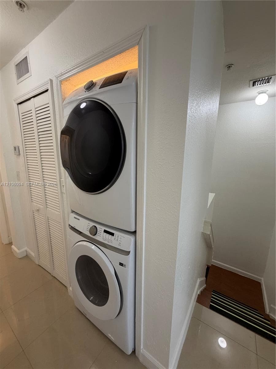 Stacked front-loading washer and dryer unit nestled in a built-in recessed wall niche featuring louvered bi-fold closet doors and neutral tile flooring