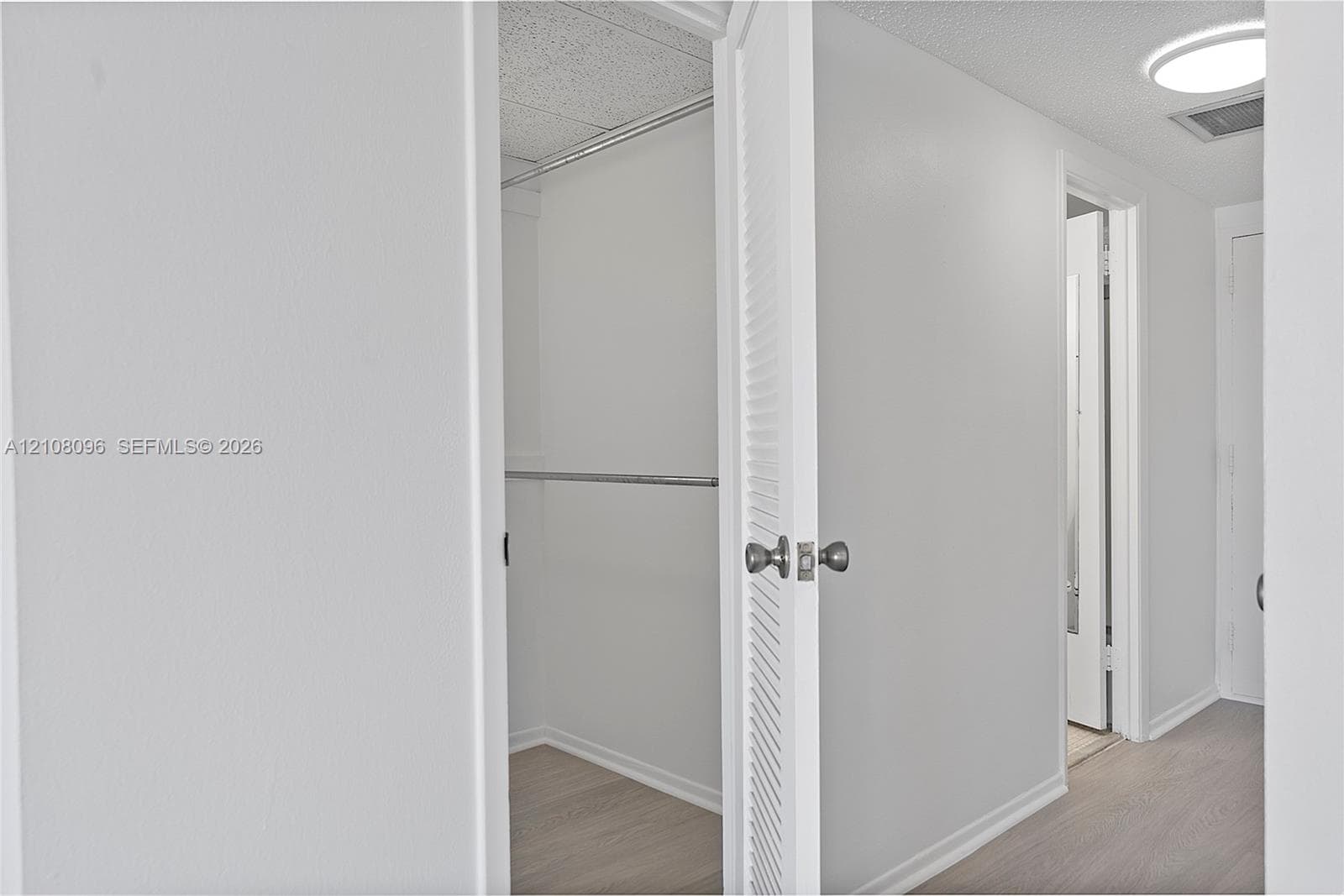 Walk-in closet featuring light-toned wood-finish flooring, white walls, and a metal hanging rod, complemented by a white louvered door with a brushed metallic knob
