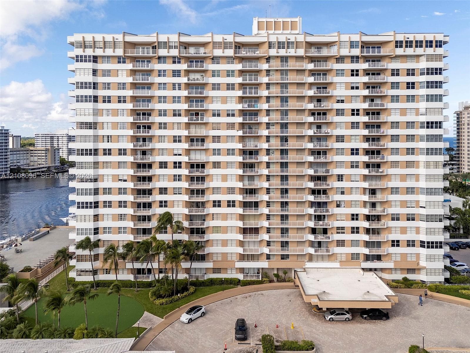 High-rise residential building featuring stacked private balconies, a covered porte-cochère, paved motor court, and landscaped grounds with mature palm trees