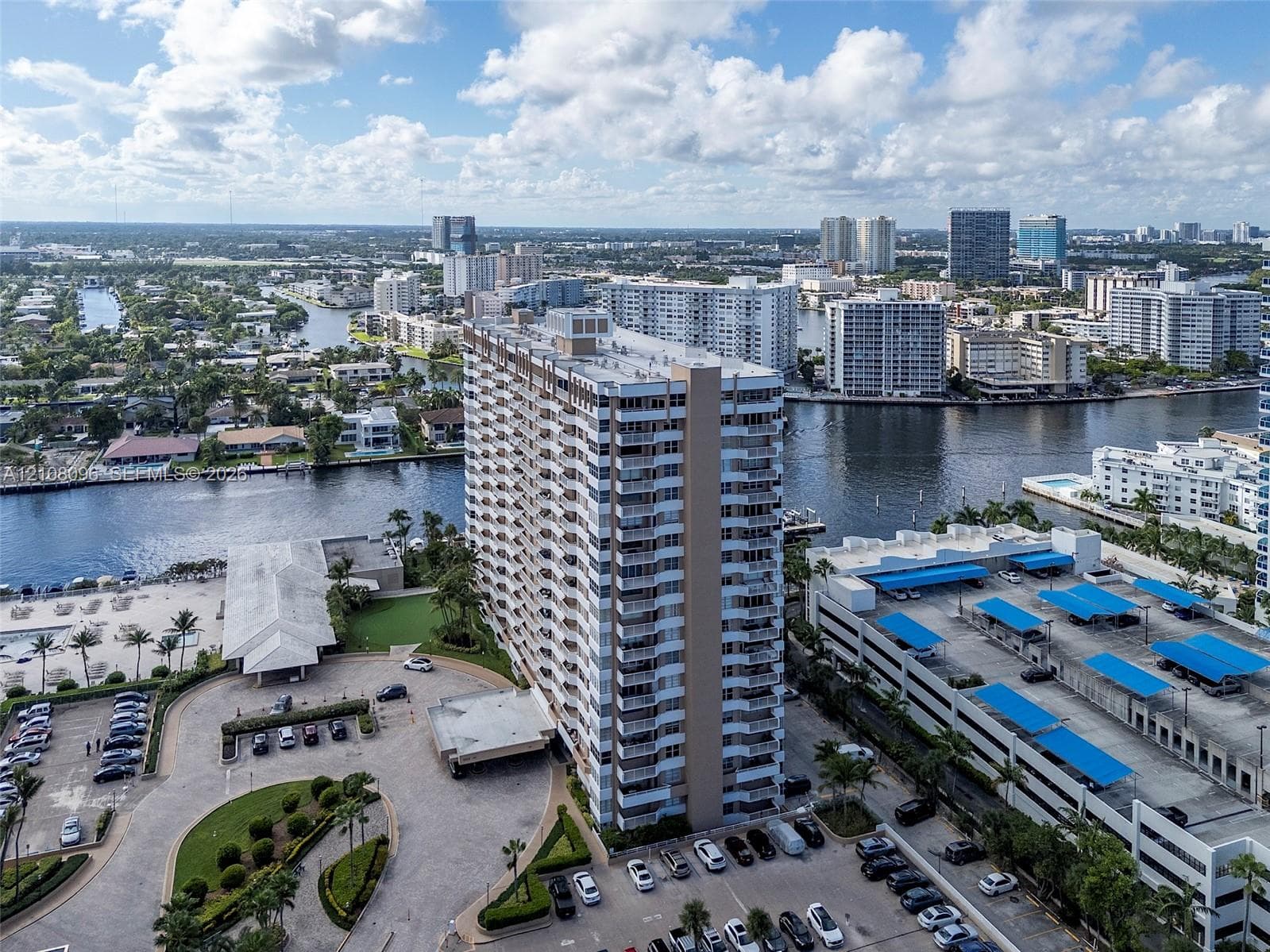 High-rise residential building featuring stacked private balconies, an attached multi-level parking structure, a paved motor court, and waterfront positioning