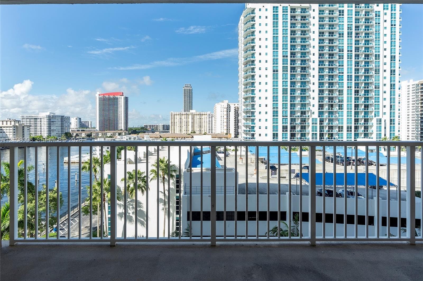 Covered balcony featuring white vertical railings and concrete flooring with urban, waterway, and palm tree scenery