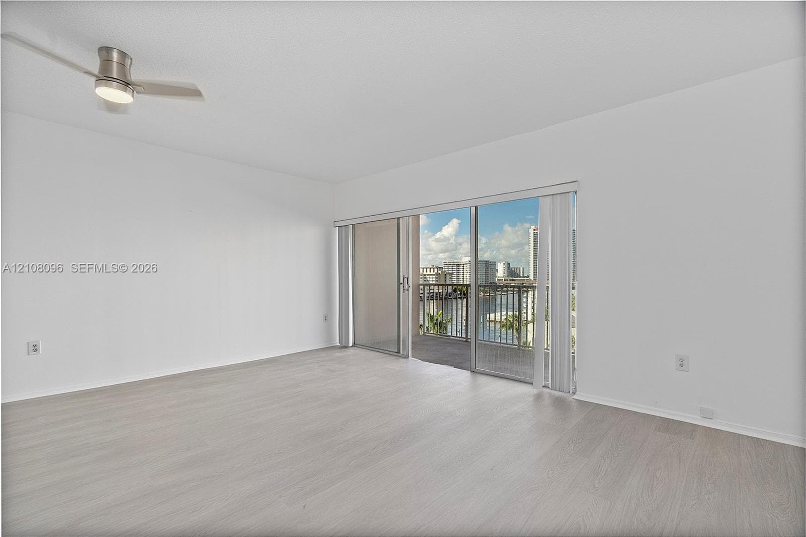Neutral interior space featuring wood-finish flooring, a ceiling-mounted light fixture with fan, and a large sliding glass door providing access to an exterior balcony