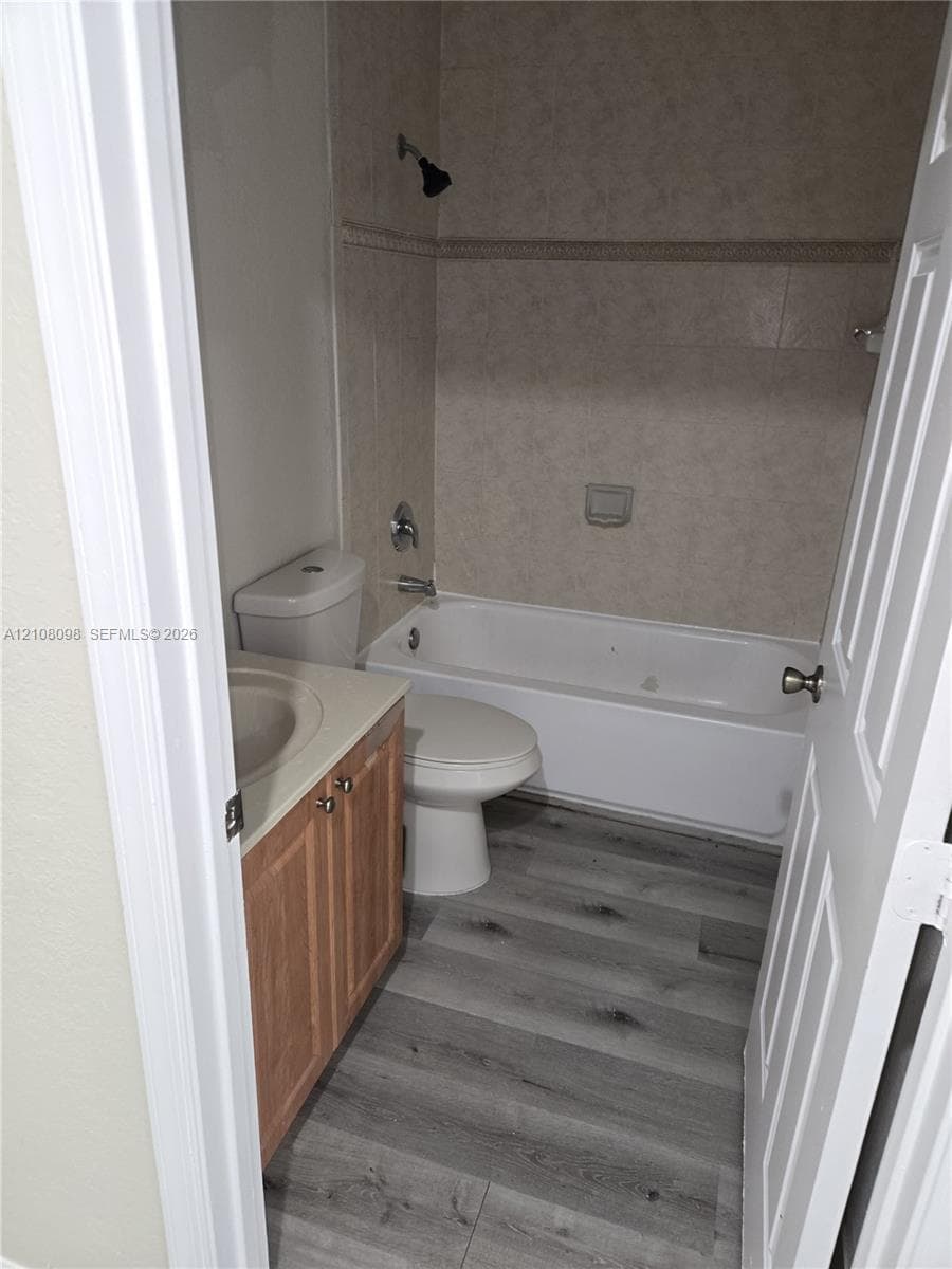 Bathroom featuring wood-finish flooring, a wood-cabinet vanity with an integrated basin, a porcelain toilet, and a tiled shower-tub combination