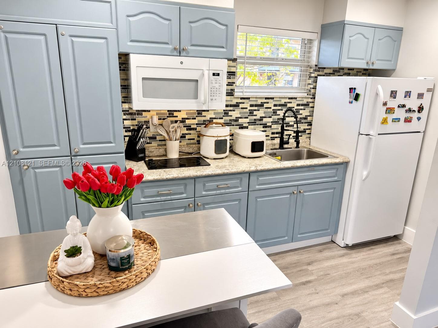 Kitchen featuring blue cabinetry, light-toned stone-finish countertops, decorative multi-tone tile backsplash, stainless steel sink with a high-arch matte black faucet, and wood-finish flooring