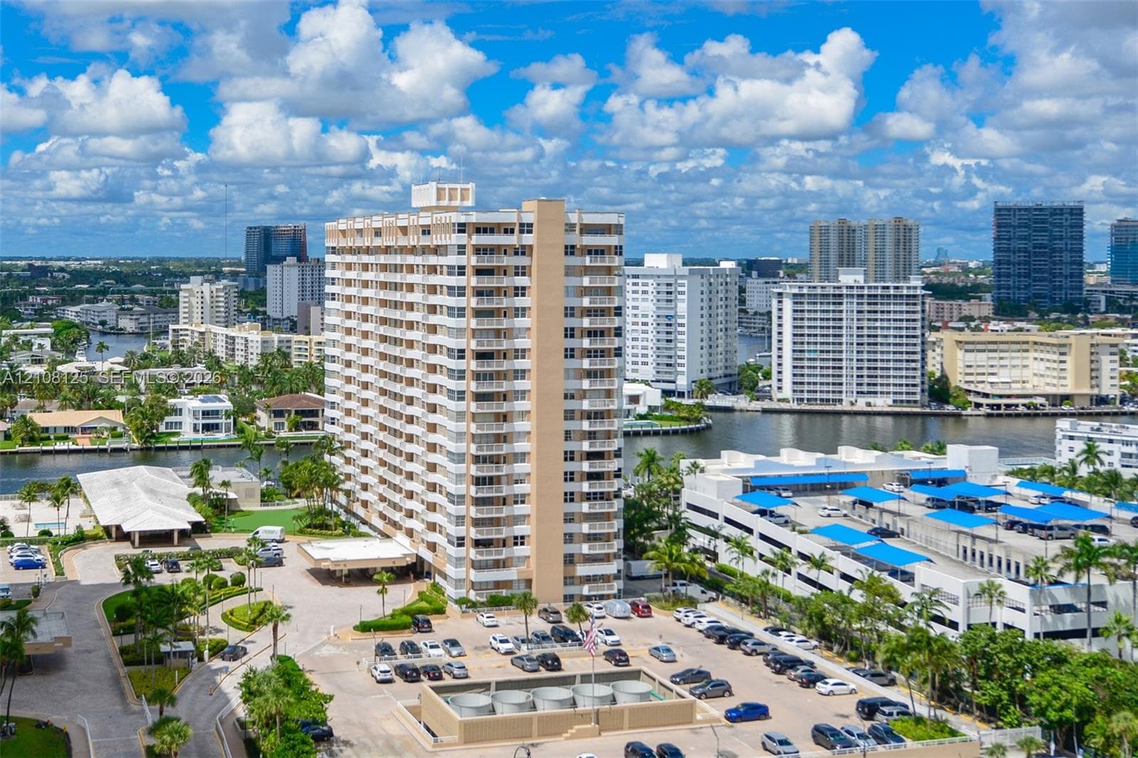 High-rise residential building featuring private balconies, expansive surface parking, a porte-cochère entry, and landscaped grounds set against a waterfront backdrop