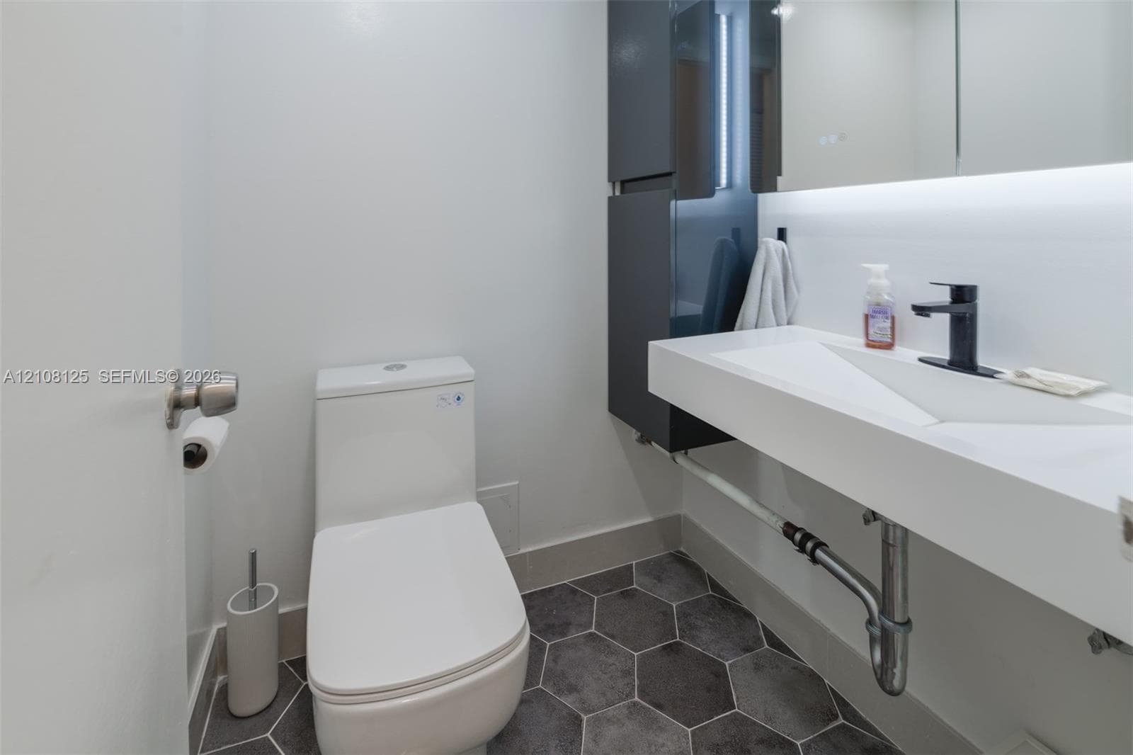 Modern powder room featuring a wall-mounted floating sink, matte black faucet, integrated storage cabinetry, back-lit mirror, and charcoal hexagon floor tile
