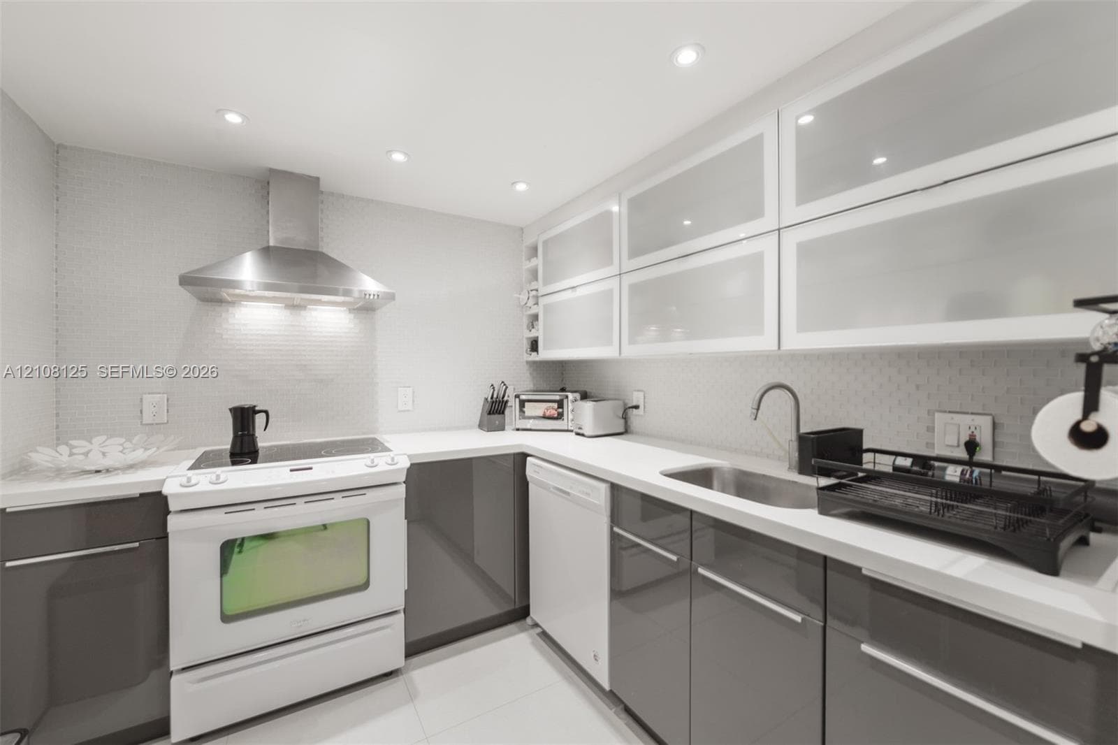 Kitchen featuring high-gloss gray lower cabinetry, white upper cabinetry with frosted glass inserts, stone countertops, stainless steel range hood, and integrated recessed lighting