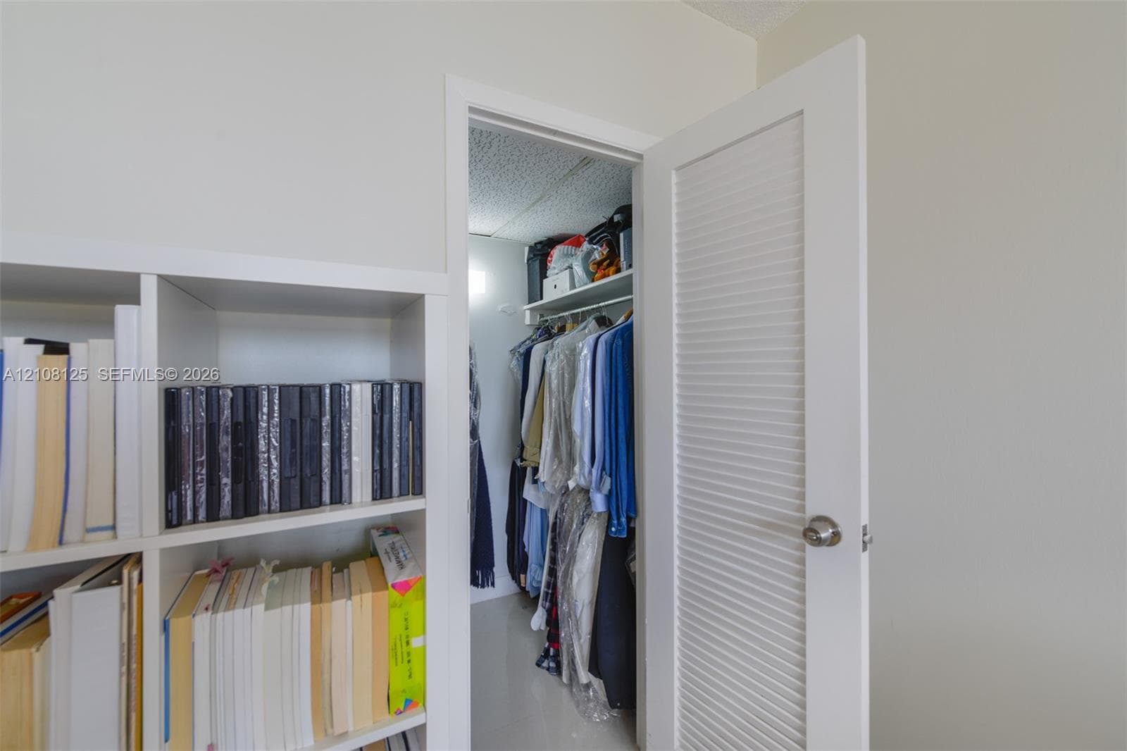 White louvered door providing entry to a storage space featuring a built-in overhead shelf and hanging rod