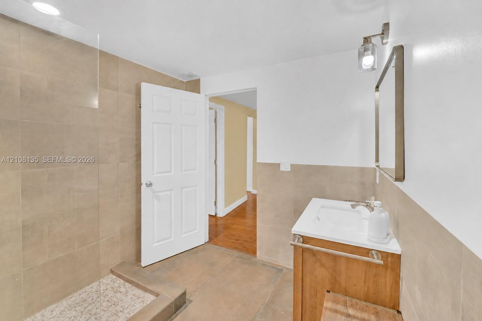 Bathroom featuring a walk-in shower with large-format tile surround, tiled shower floor, glass partition, wood-finish vanity, and wall-mounted lighting