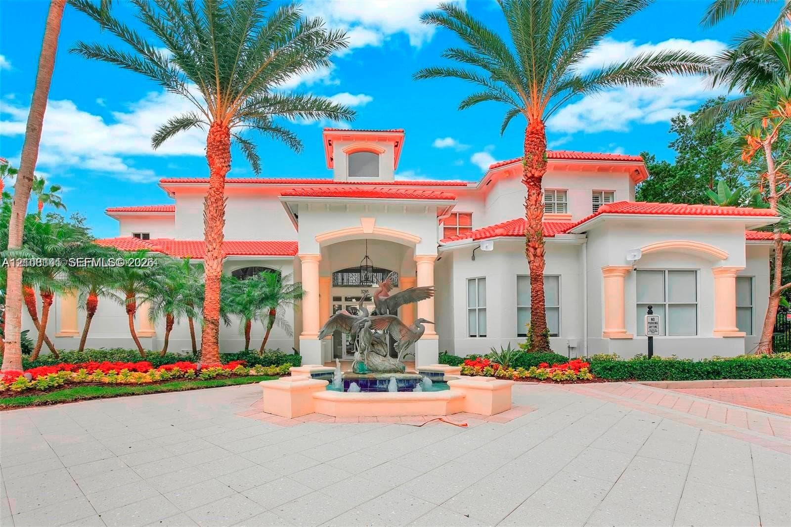 Mediterranean-style exterior featuring a red tile roof, stucco facade, covered entryway with columns, a central decorative fountain, and palm-lined landscaping