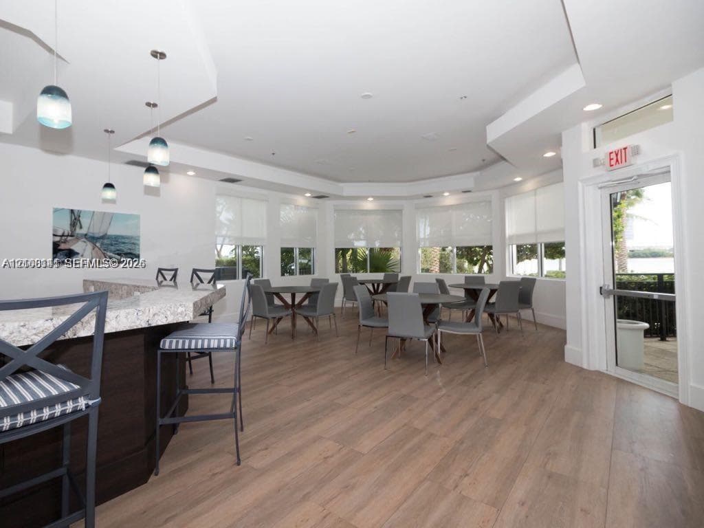 Multipurpose room featuring wood-finish flooring, a built-in bar with stone countertop, multi-tier recessed ceiling, and floor-to-ceiling windows with roller shades