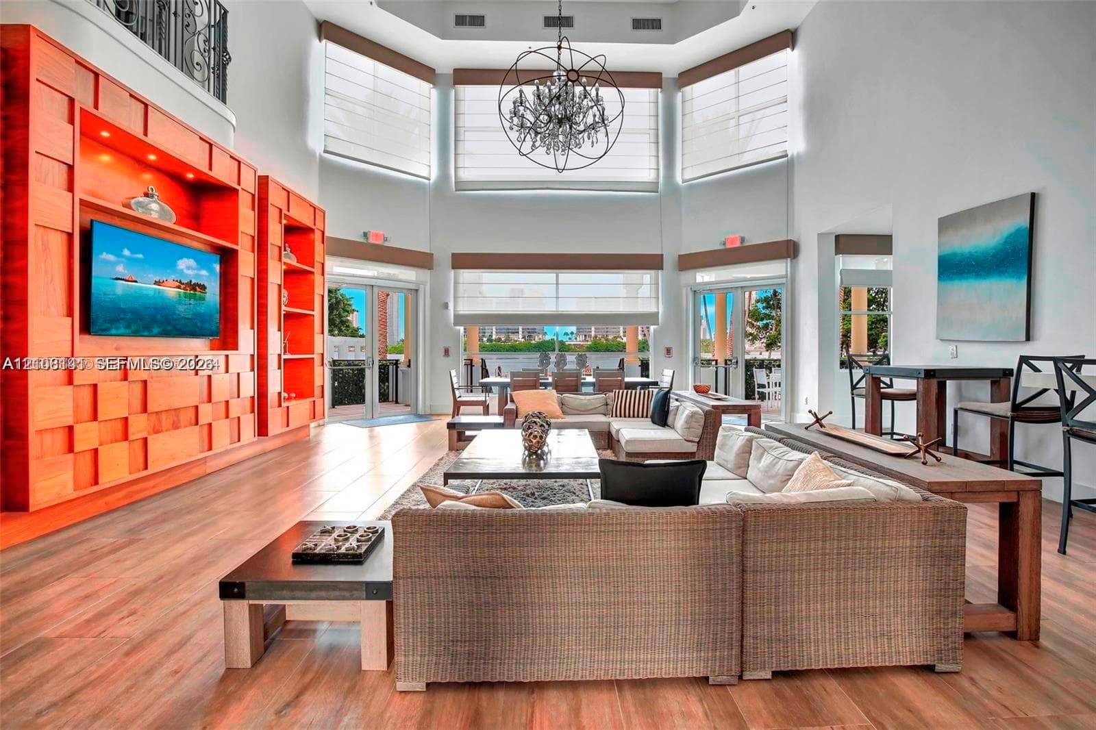 Double-height ceilings with large multi-panel windows, custom floor-to-ceiling wood-finish media cabinetry, wood-finish flooring, and a large decorative chandelier