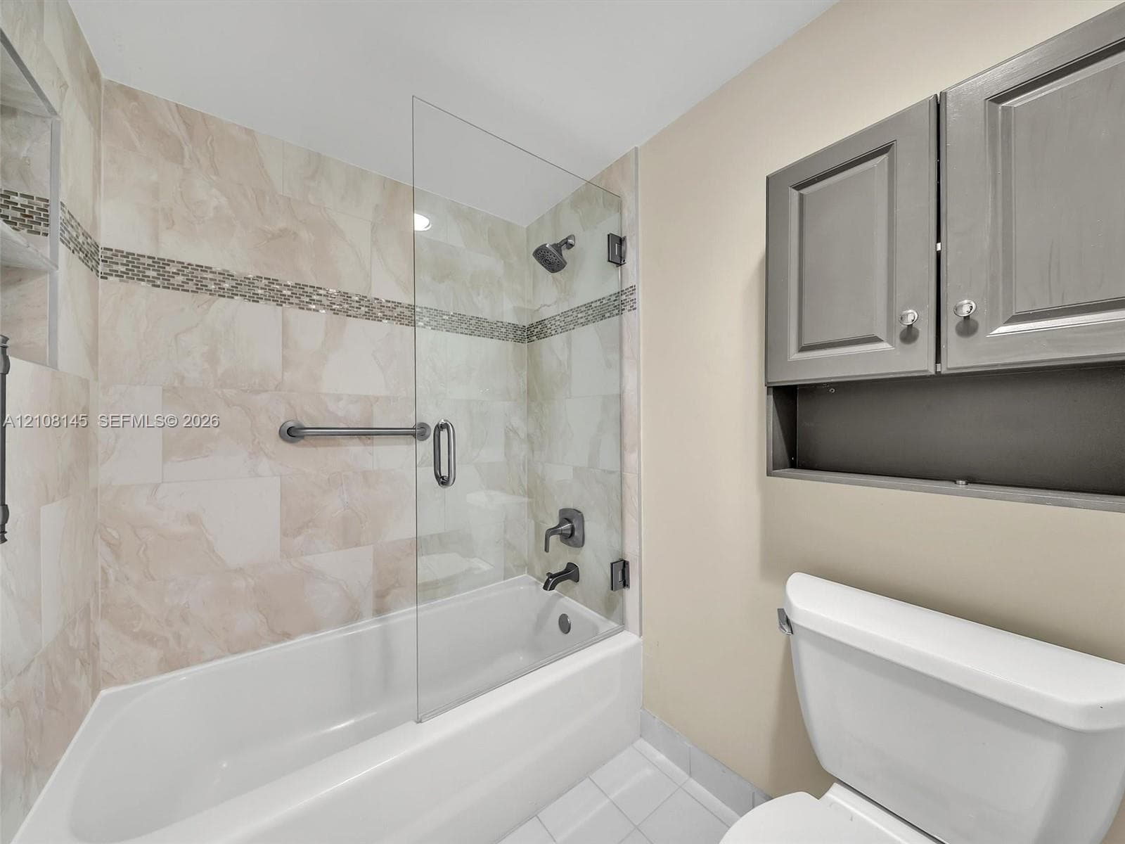 Bathtub featuring a glass shower panel, neutral tile surround with decorative mosaic accent, matte black wall-mounted fixtures, and built-in grey cabinetry