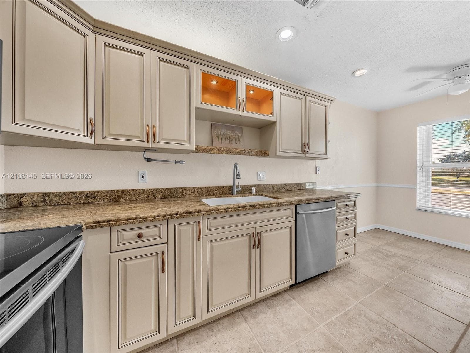 Kitchen featuring light-toned raised-panel cabinetry, stone-finish countertops, stainless steel dishwasher, integrated glass-front display shelving, and neutral tile flooring