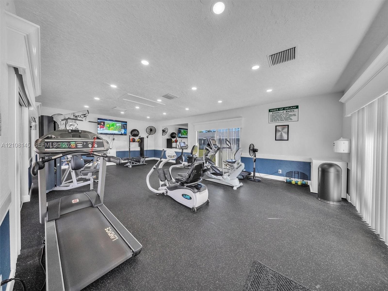 Fitness area featuring rubberized flooring, recessed ceiling lighting, wall-mounted mirrors, and vertical blinds