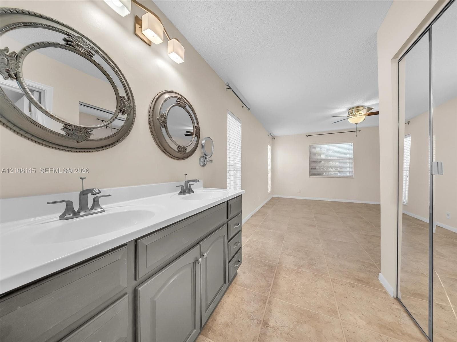 Dual-sink vanity with gray cabinetry and white countertop, ceramic tile flooring, ceiling fan, and mirrored closet doors