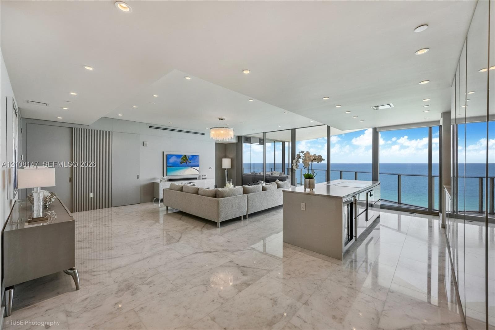 Open-concept living space featuring polished stone flooring, floor-to-ceiling glass wall panels, an integrated kitchen island with stone countertop, recessed ceiling lighting, and vertical-slat wall paneling