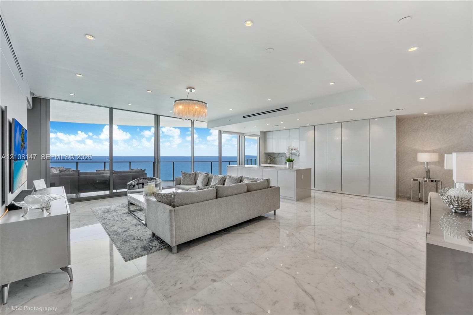 Open-concept interior featuring floor-to-ceiling glass panel walls, light-toned stone-finish flooring, recessed lighting, sleek high-gloss cabinetry, and an integrated kitchen island