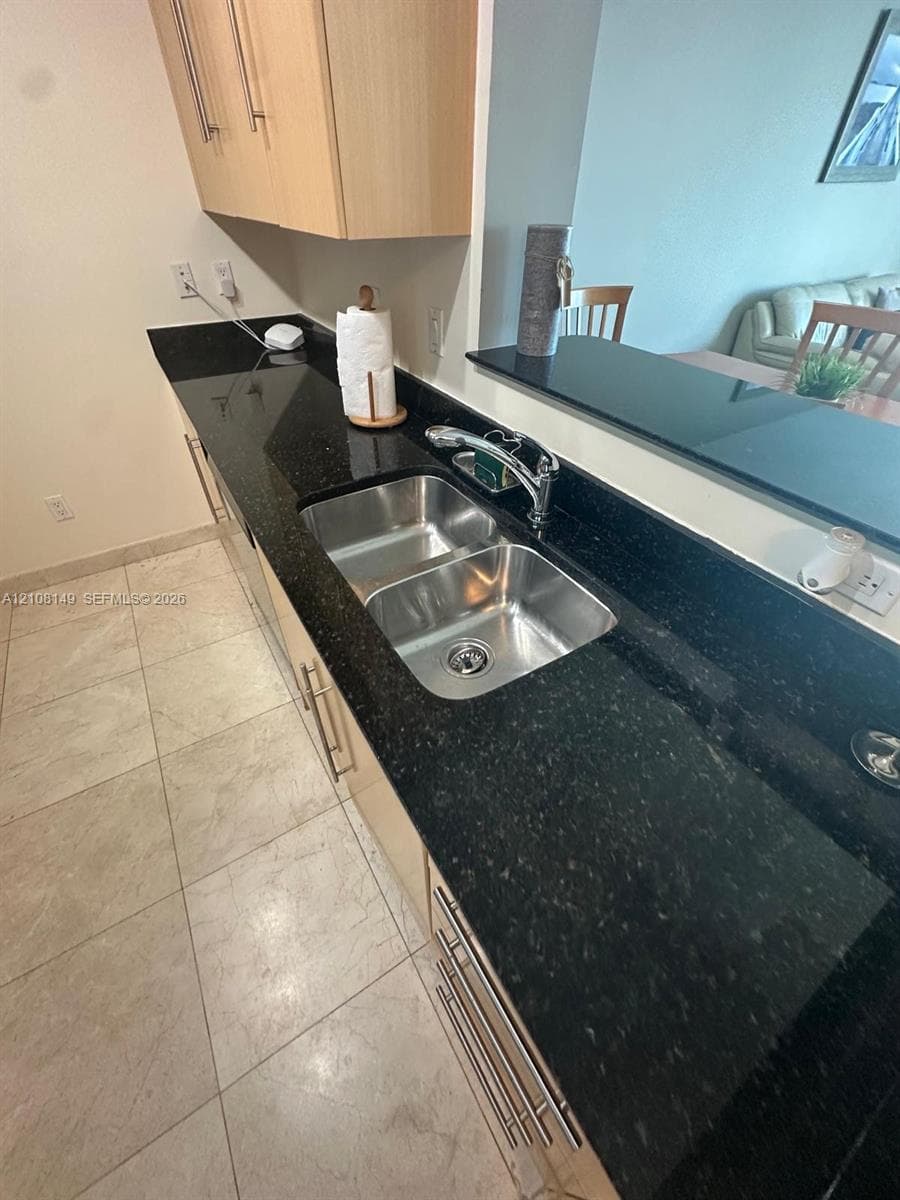 Kitchen featuring dark stone countertops, light wood-finish cabinetry with metallic hardware, dual-basin stainless steel sink, polished chrome faucet, and tile flooring