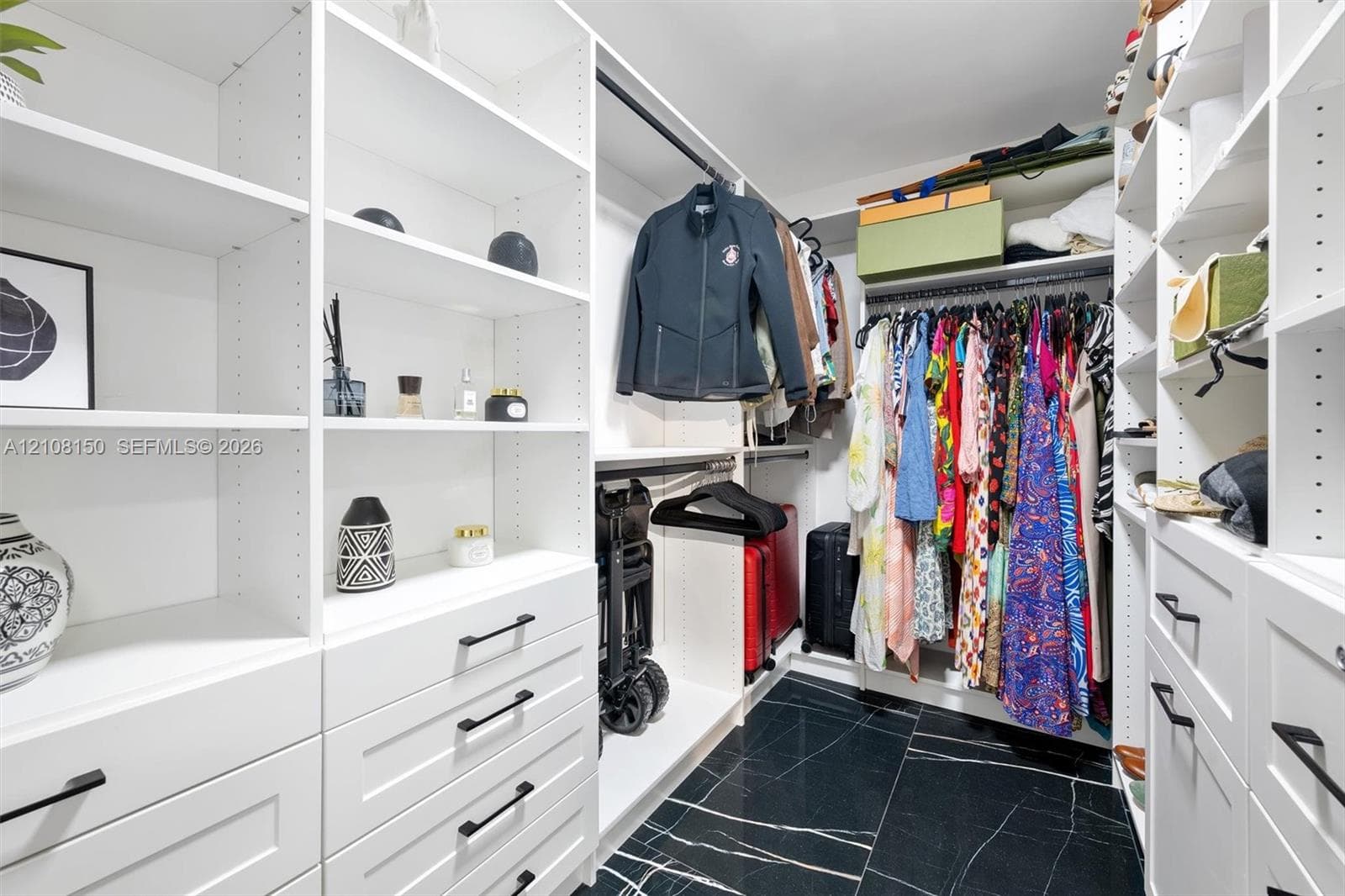 Custom walk-in closet featuring white built-in shelving, floor-to-ceiling storage units, multiple drawers with matte black hardware, integrated hanging rods, and dark stone-finish tile flooring