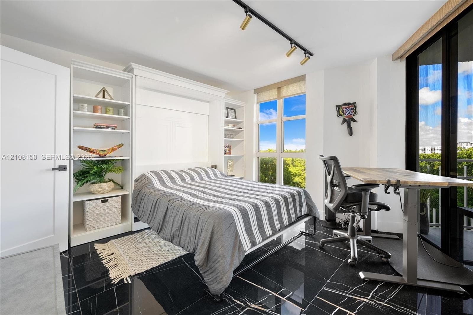 Built-in floor-to-ceiling white shelving, integrated wall bed system, black tile flooring with veining, track lighting, and full-height windows