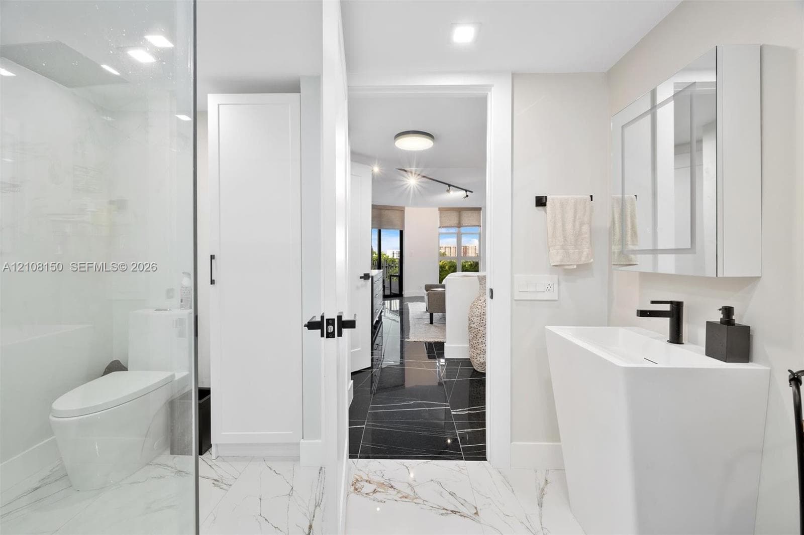 Modern bathroom featuring a tall white pedestal sink with matte black hardware, a glass-enclosed shower, built-in cabinetry, and white tile flooring with veining