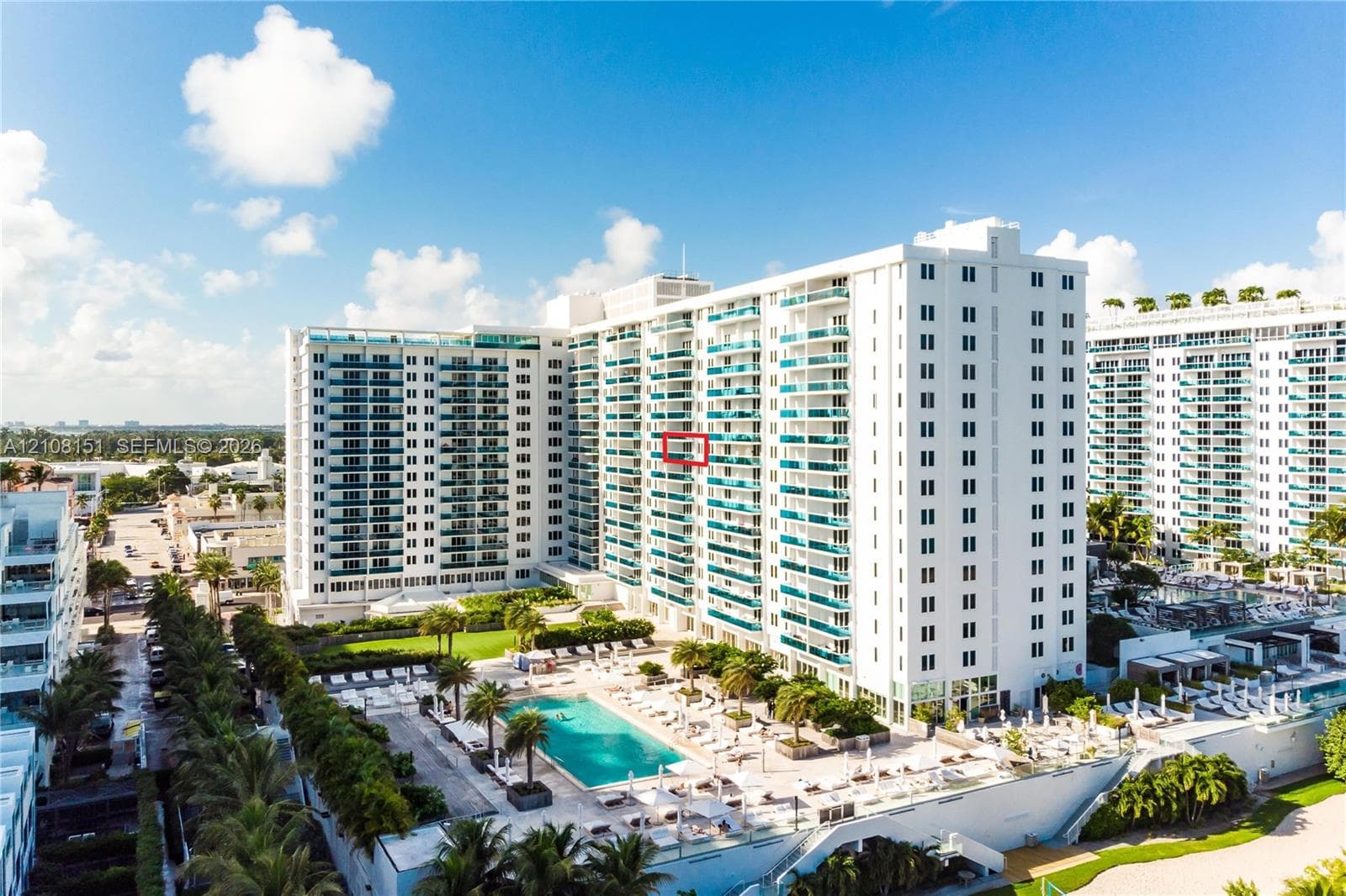 High-rise residential building featuring expansive private balconies with glass railings, an outdoor swimming pool, manicured lawn areas, and mature palm trees