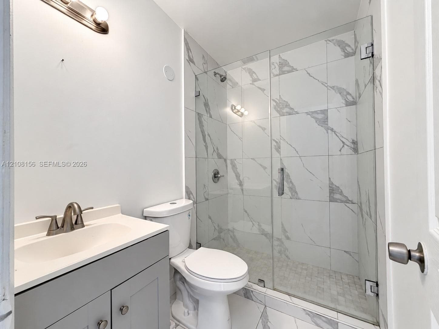 Bathroom featuring a grey shaker-style vanity with an integrated sink, walk-in shower with large-format stone-patterned tile, frameless glass enclosure, and modern brushed-metal fixtures