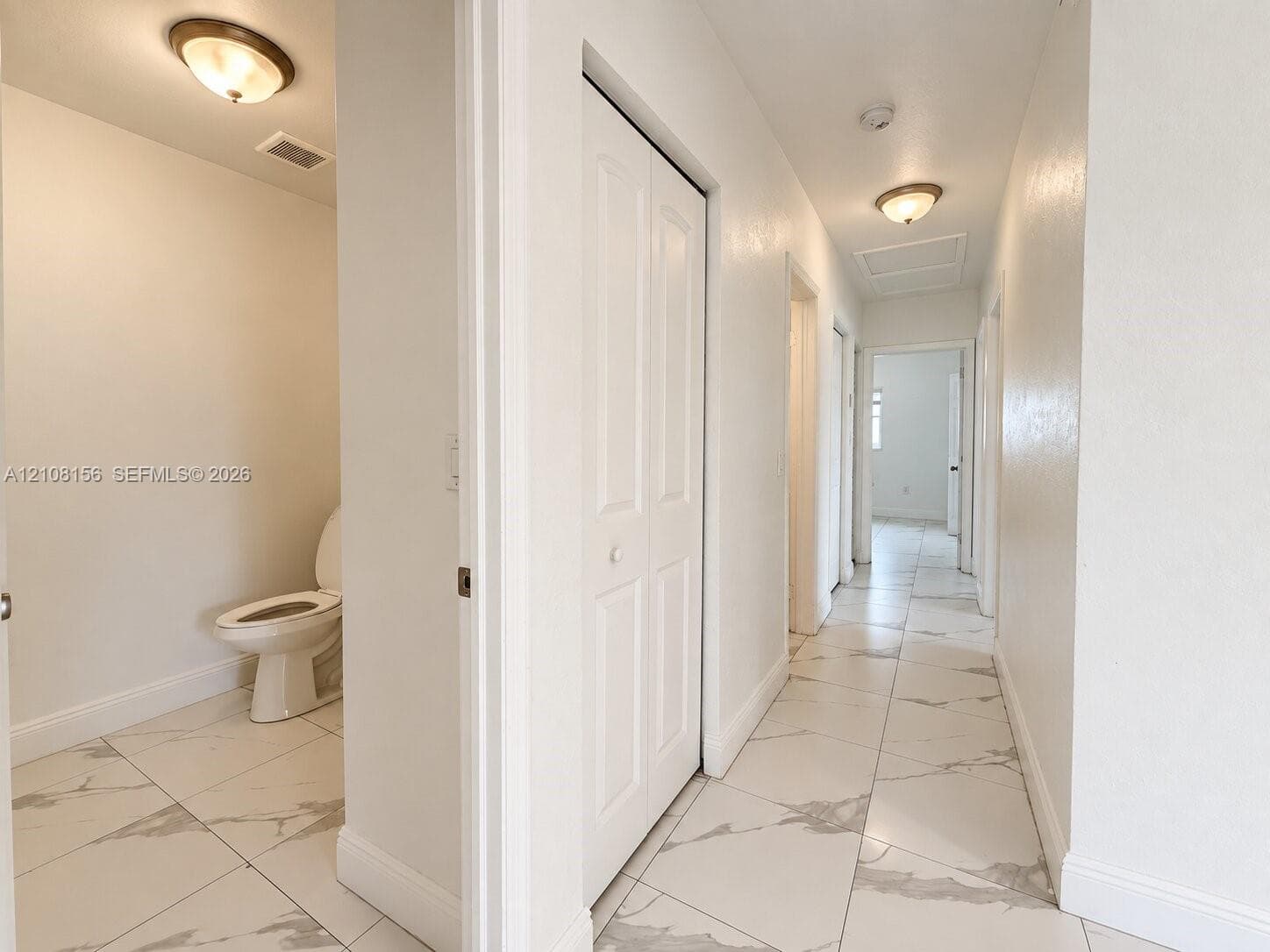 Hallway featuring large-format white stone-finish floor tiles with grey veining, crisp neutral wall paint, white bi-fold closet doors, and flush-mount lighting fixtures
