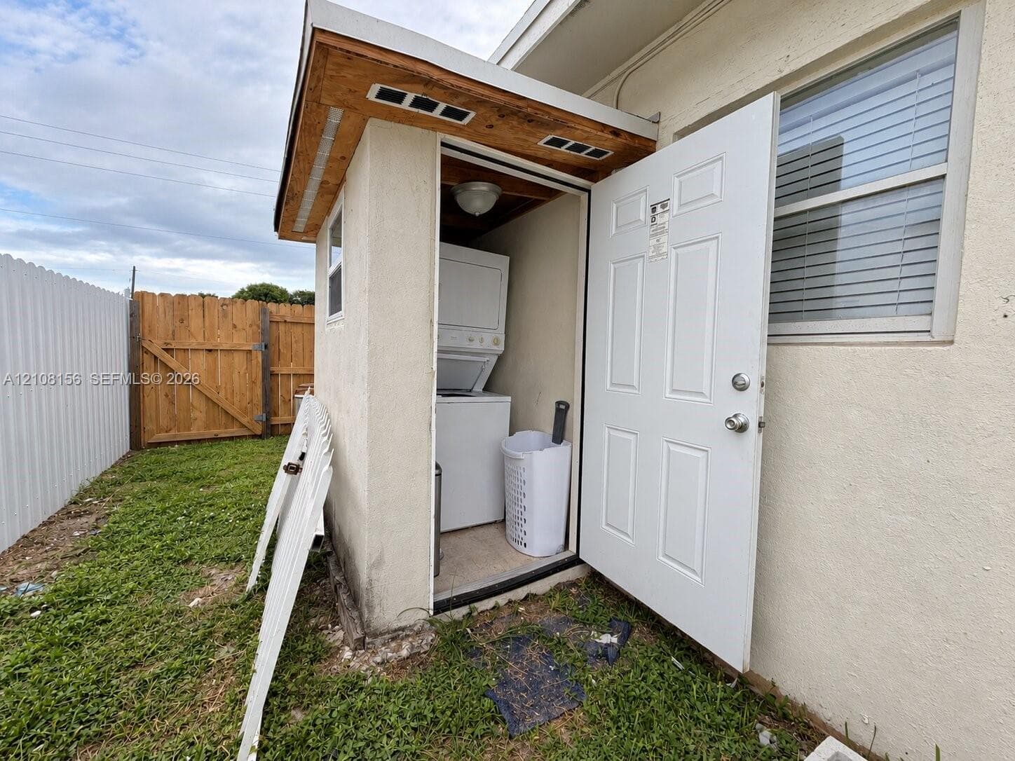 Exterior utility enclosure featuring a stucco finish, a shingled shed-style roof, a single-entry door, and stacked laundry appliances