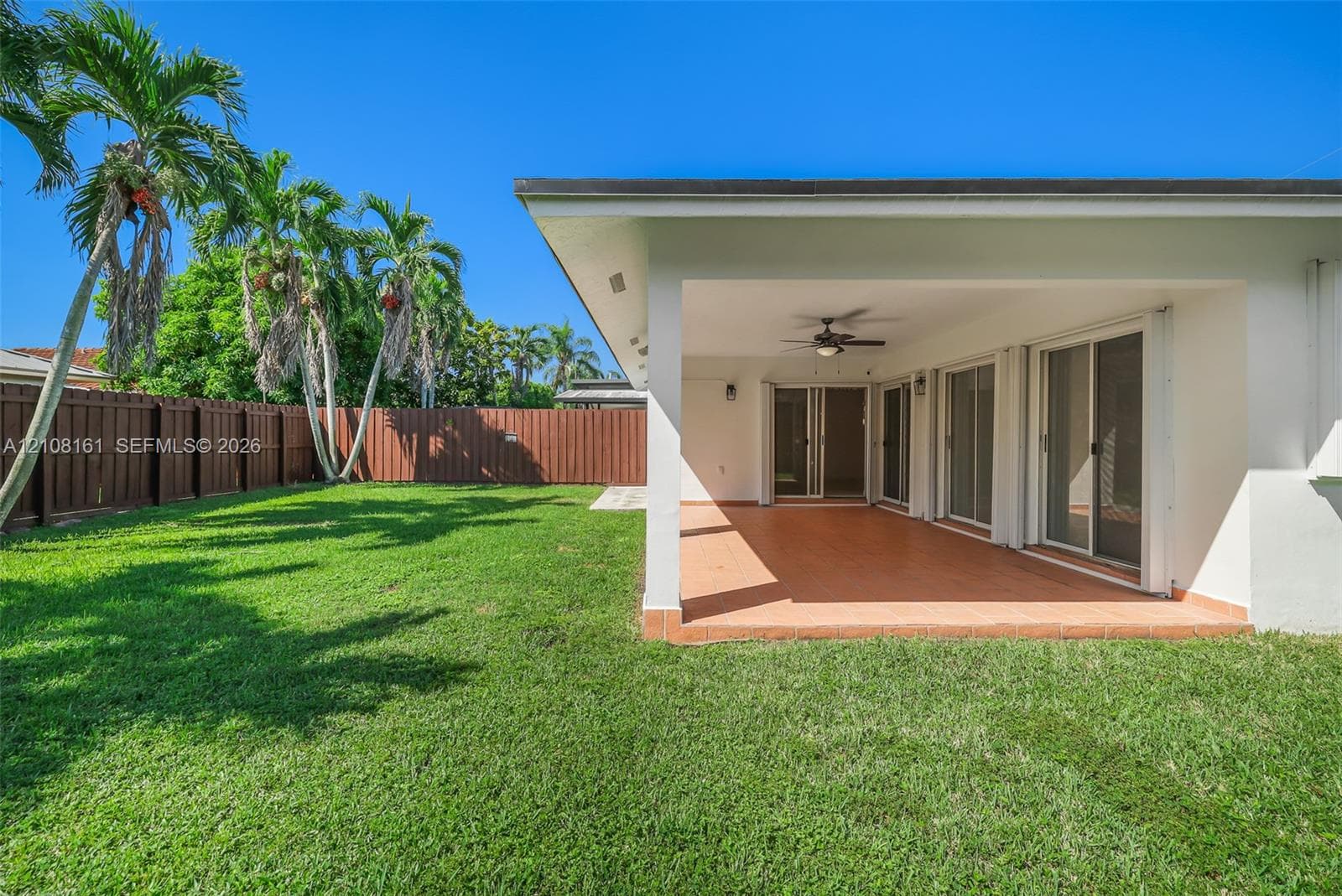 Covered concrete patio with a ceiling fan, sliding glass door access, privacy wood fencing, and mature palm trees