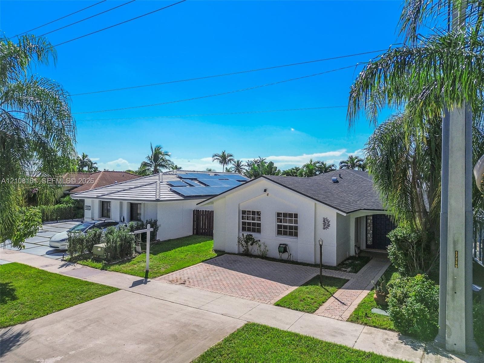 Single-story exterior featuring white stucco siding, a shingled roof with integrated solar panels, paved brick driveway, and mature landscaping