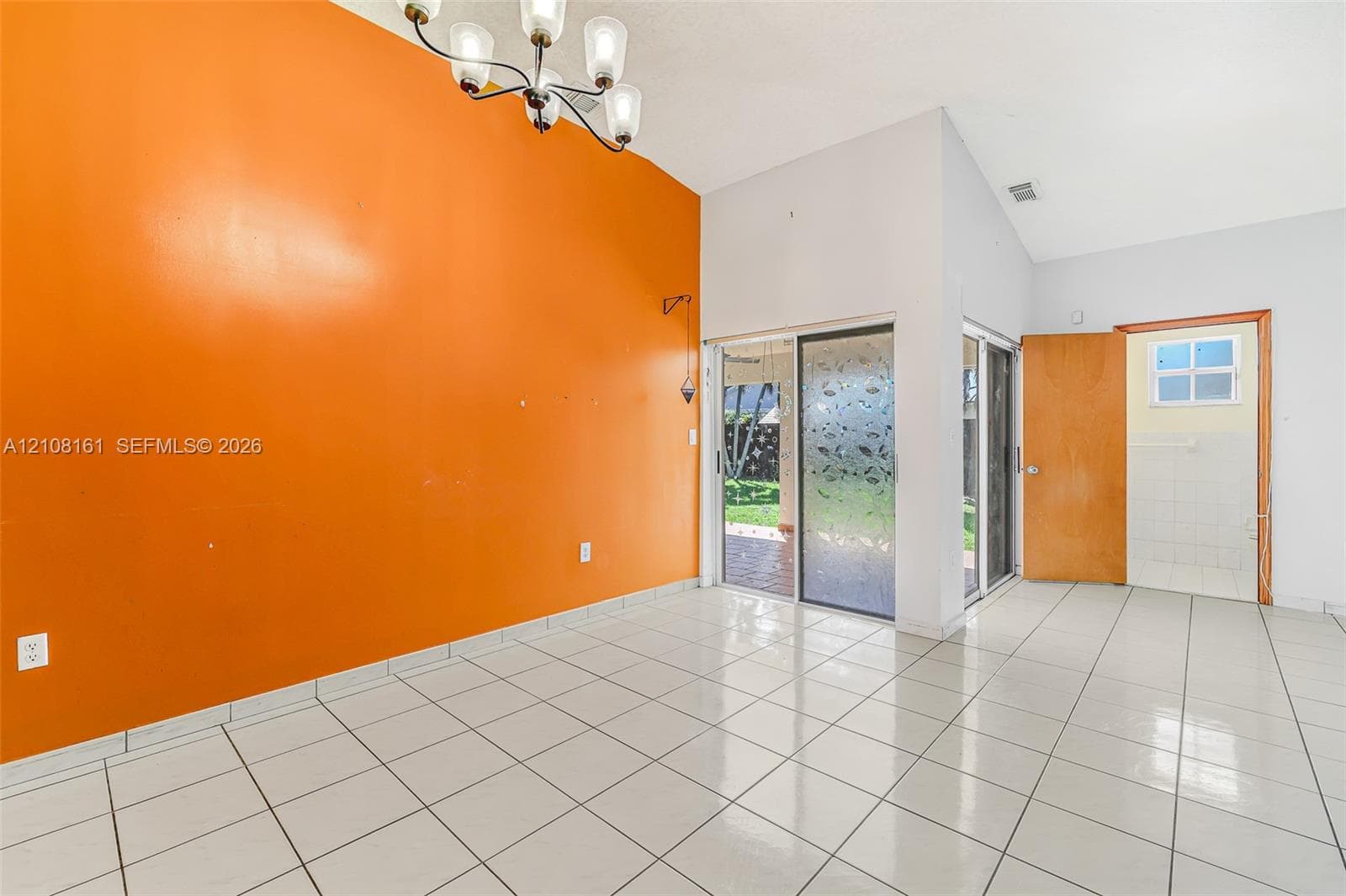 High-ceiling interior space featuring large-format ceramic tile flooring, a multi-arm chandelier, sliding glass doors with frosted glass detailing, and a solid wood entry door