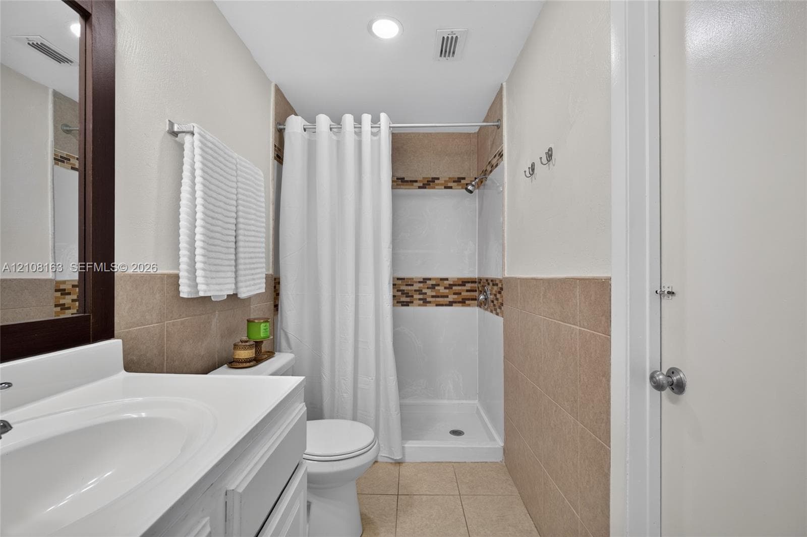 Bathroom featuring a white vanity with a porcelain sink, beige wall tiling, mosaic tile accent border, and a walk-in shower with white surround