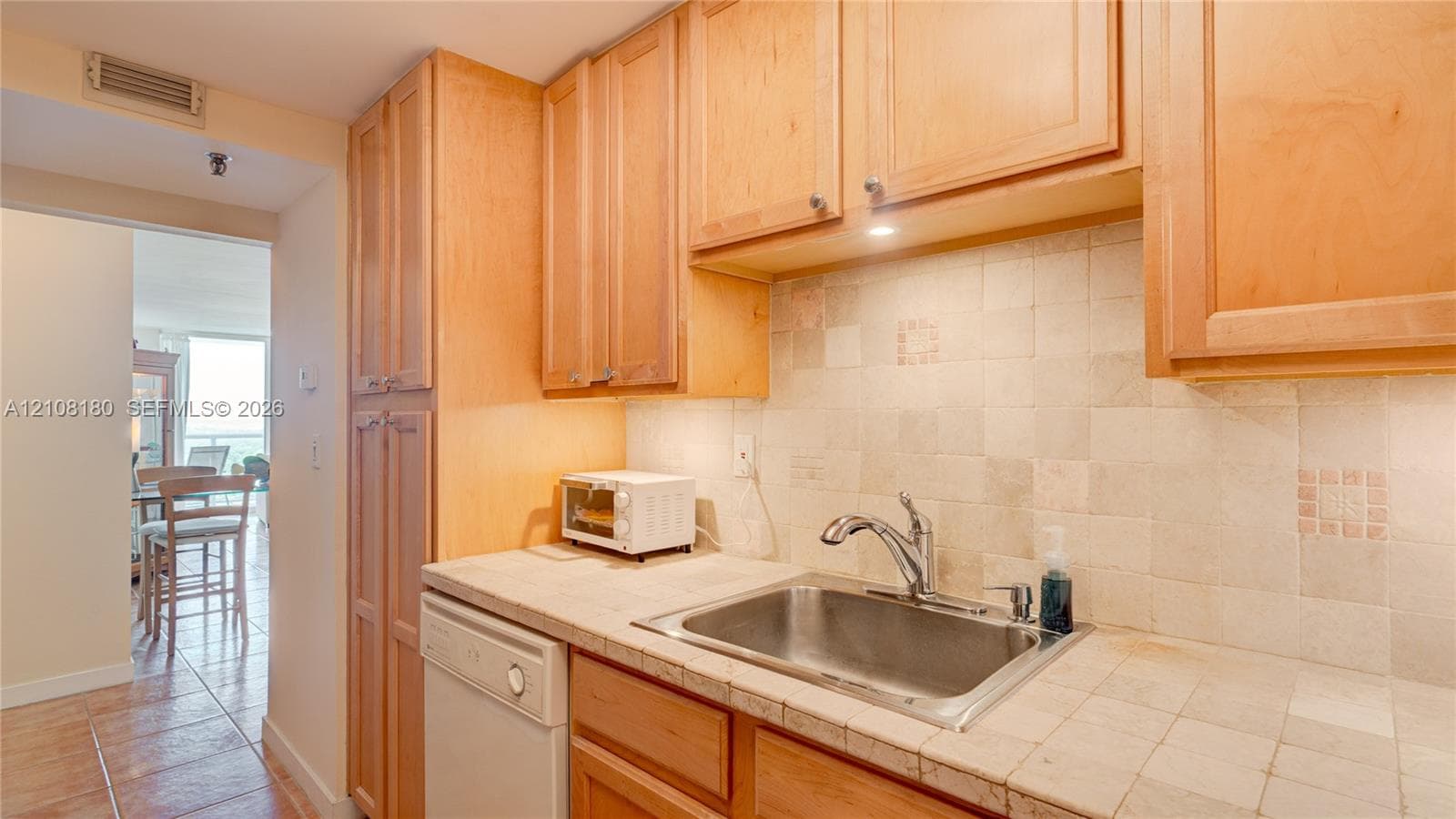 Kitchen featuring light-finish wood cabinetry, tile countertops, matching tile backsplash, and a stainless steel sink with a high-arch faucet