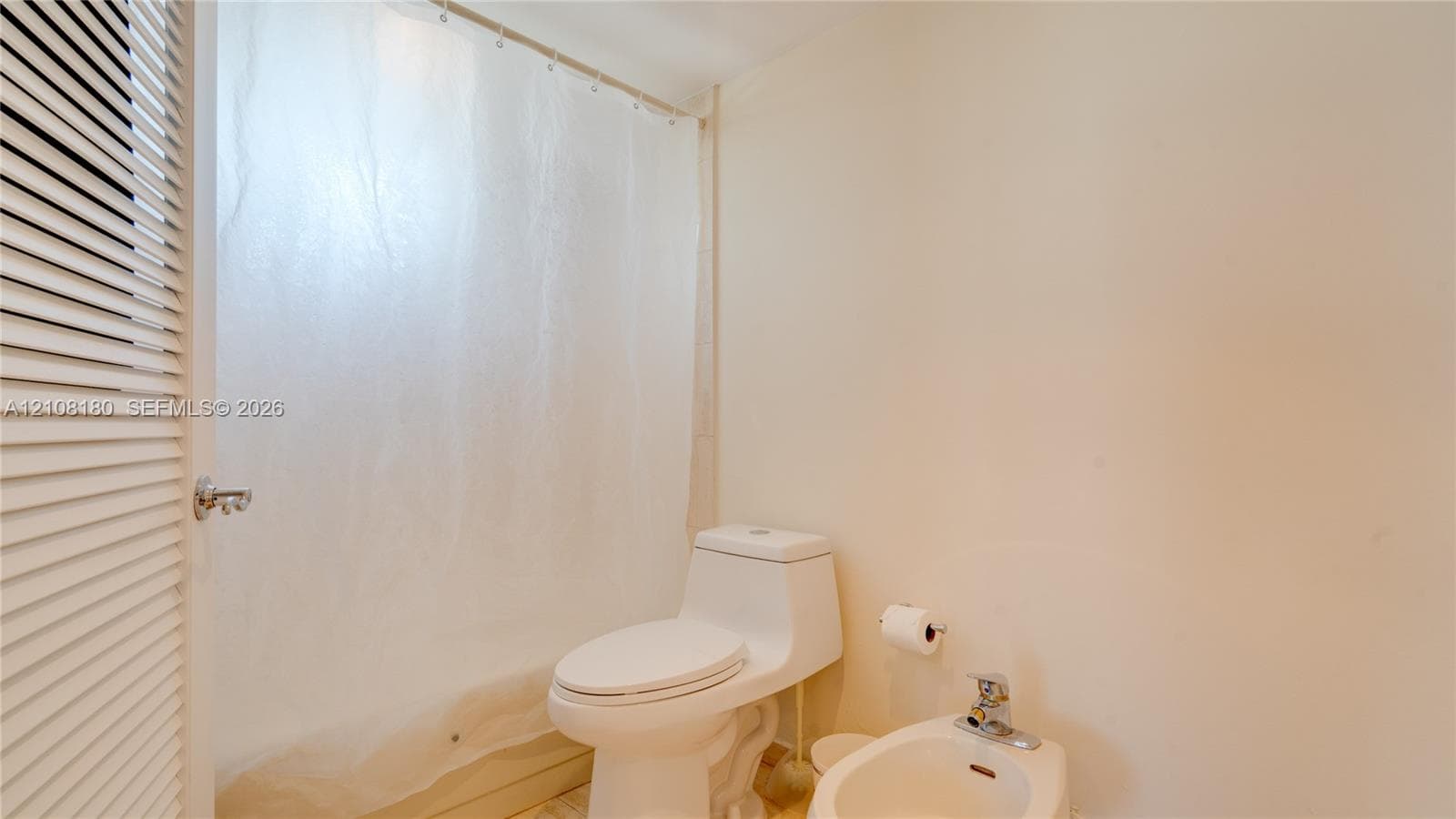 Bathroom featuring a white elongated toilet, porcelain bidet with chrome faucet, shower-tub combination with a curtain, and louvered closet door