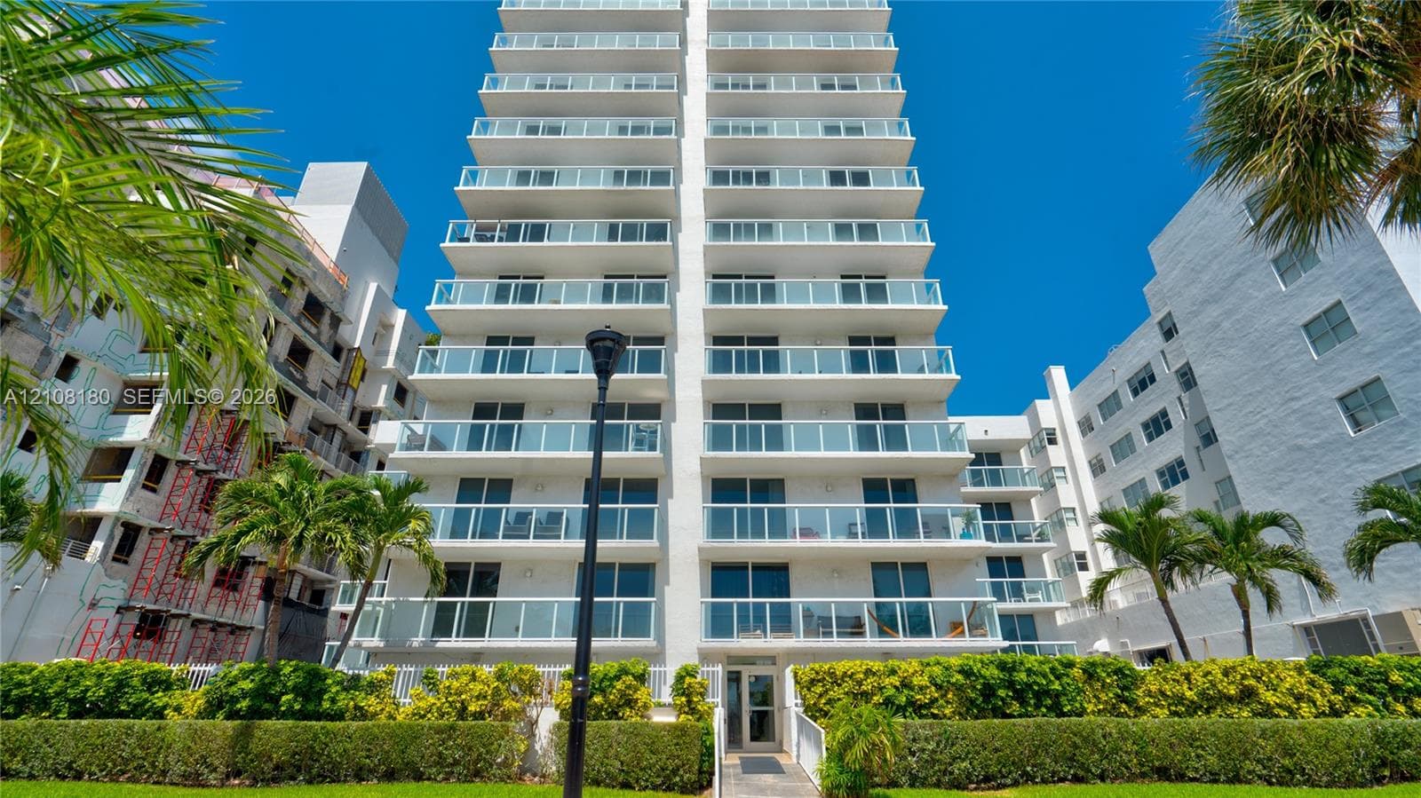 Multi-story high-rise exterior featuring stacked glass-railing balconies, floor-to-ceiling windows, white facade, and manicured landscaping