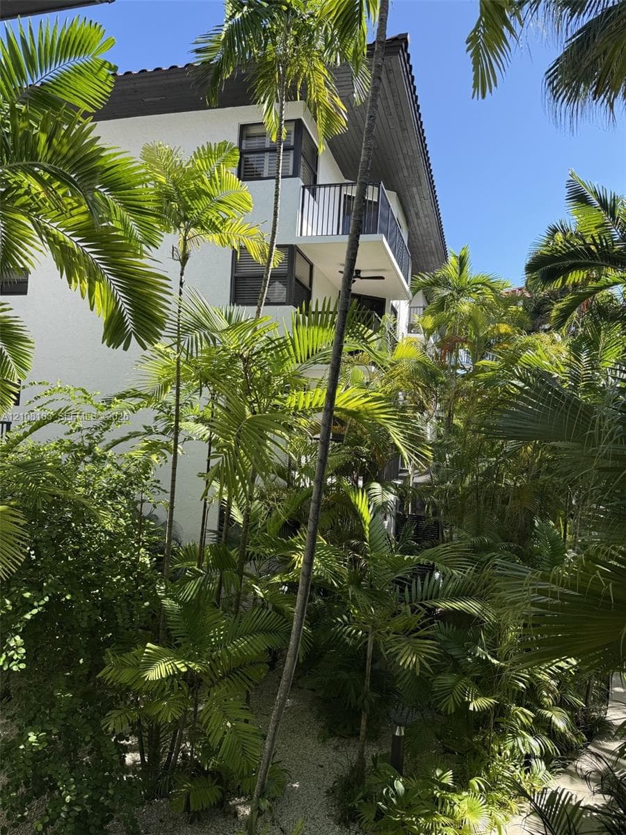 Multi-story exterior featuring white stucco siding, a tiled roof, metal balcony railings, and lush palm landscaping