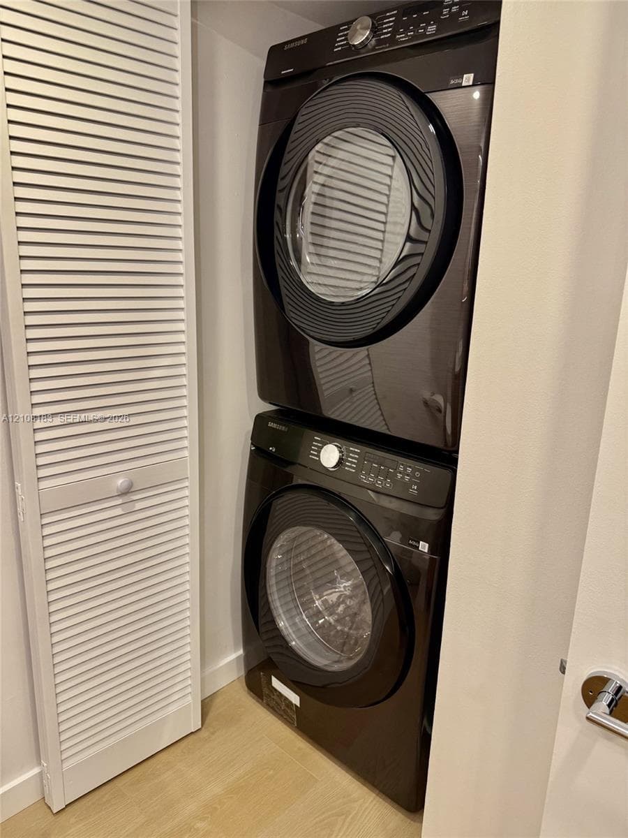 Stacked black high-efficiency laundry appliances set within a dedicated closet featuring louvered bi-fold doors and wood-finish flooring