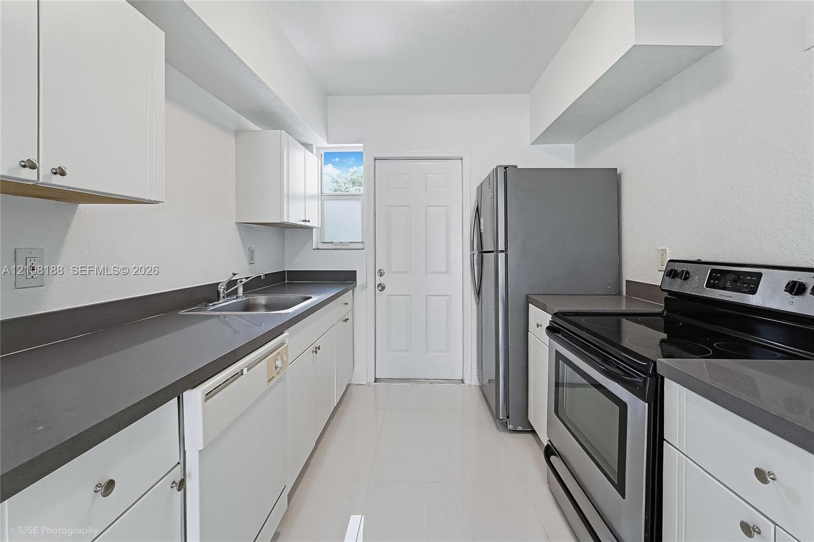 Galley kitchen featuring white shaker-style cabinetry, solid-surface countertops, stainless steel appliances, a recessed stainless steel sink with a pull-out faucet, and tile flooring