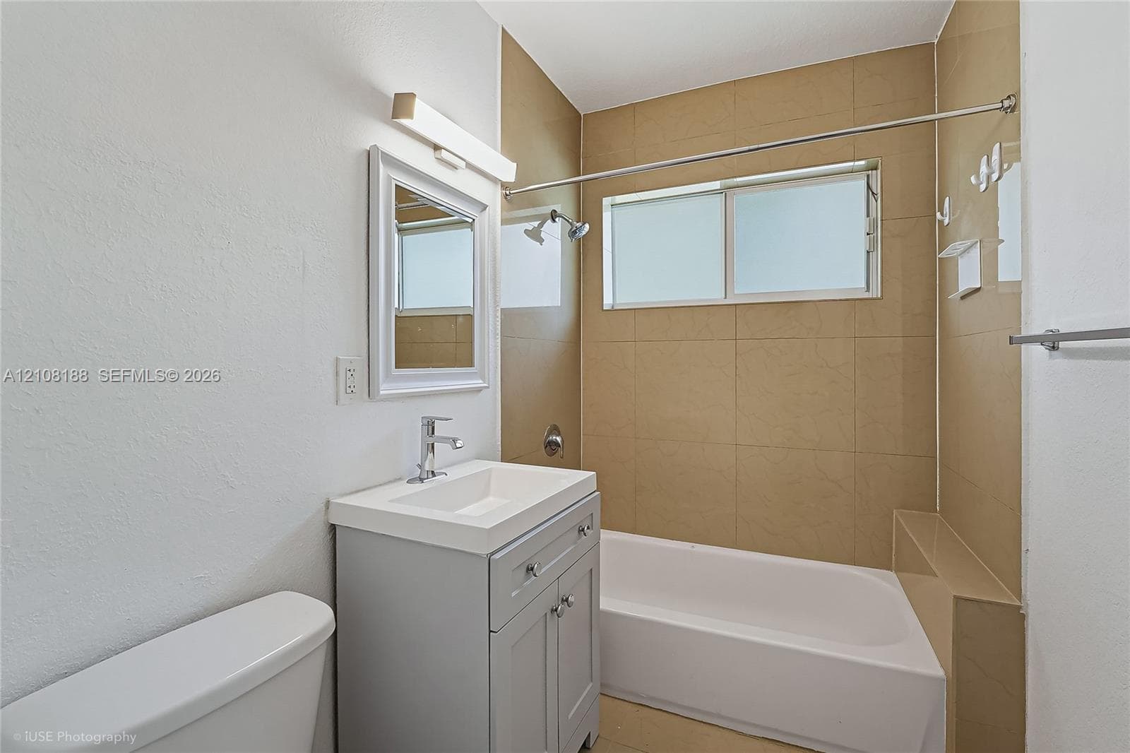 Bathroom featuring a grey vanity with a rectangular sink, brushed nickel faucet, wall-mounted vanity light, and a tub-shower combination with floor-to-ceiling neutral tile surround