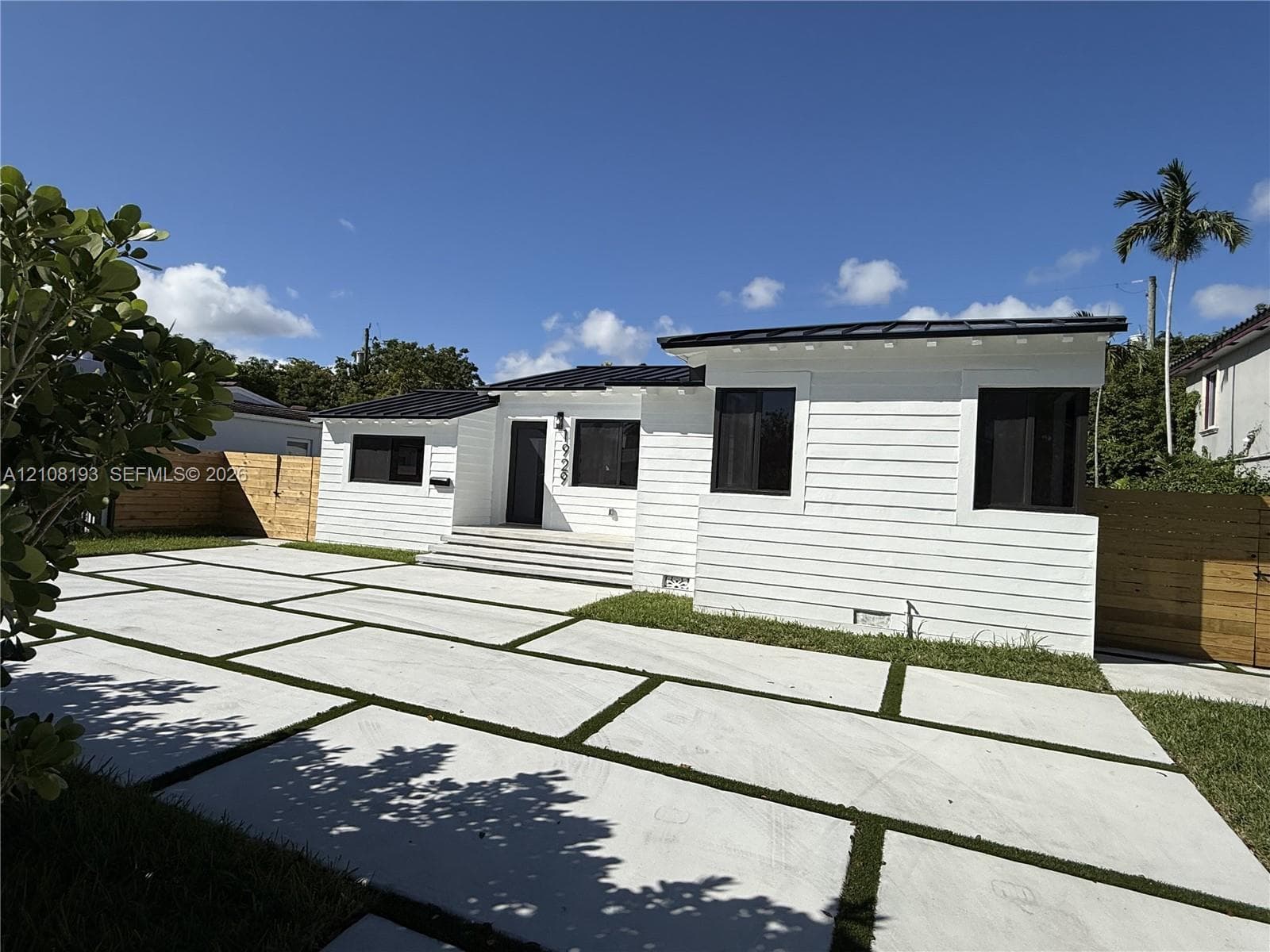 Single-story residence featuring white horizontal siding, a dark metal roof, geometric concrete paver driveway with turf accents, and wood-paneled privacy fencing